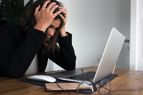 Female holding her head with both hands, looking at a laptop on a desk.