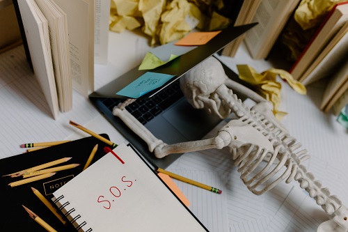 A skeleton with its head down and arms up is lying on the laptop surrounded by textbooks and paper. “SOS” is written on the notebook to the left of the laptop.