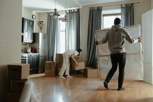 An open floor plan of a living space with a kitchen and 2 large windows with grey drapery. The room is surrounded by boxes. A female is stacking a box onto another. A male is carrying a large picture to the corner near the door.