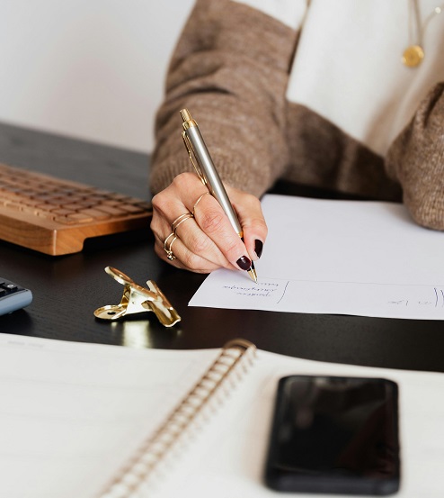 Female writing on a piece of paper with a keyboard and a cell phone nearby.