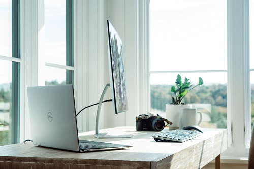 A laptop, monitor, keyboard, camera and a plant is on a white desk that is in front of a set of windows.