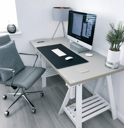 An office chair is in front of a desk with a computer screen, a lamp and a small plant.